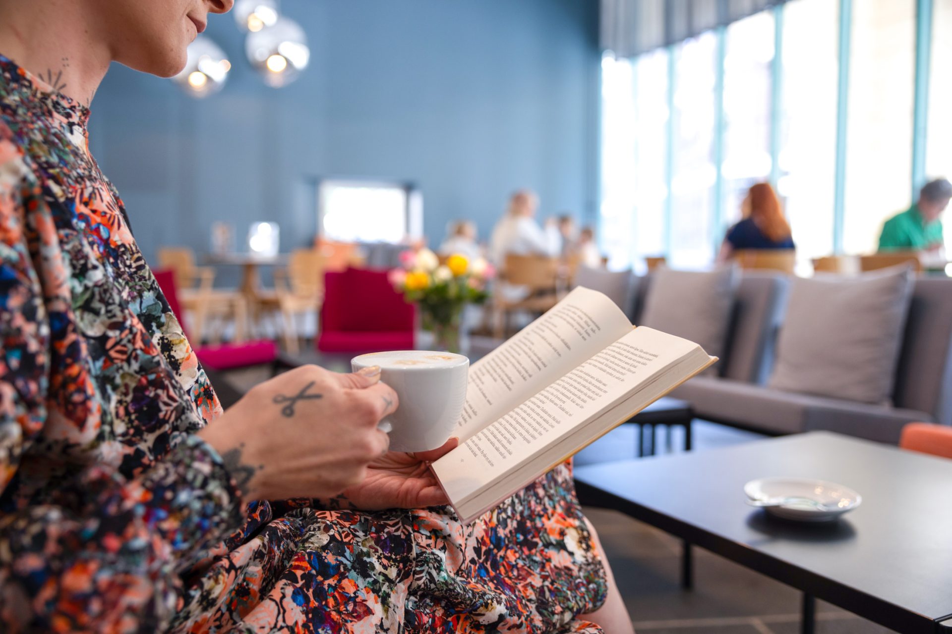 Nainen lukee kahvilassa kirjaa kahvikuppi toisessa kädessä