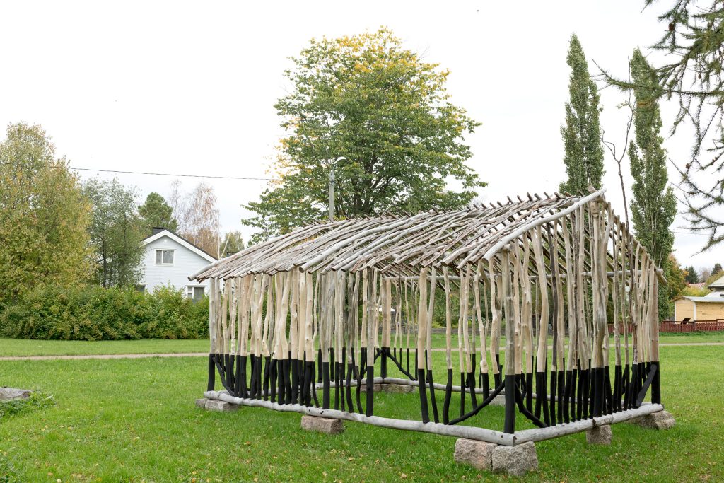 Puumateriaalista paikalla rakennettu teos puistossa, teoksen muoto viittaa pientalovaltaiseen sijaintiympäristöön