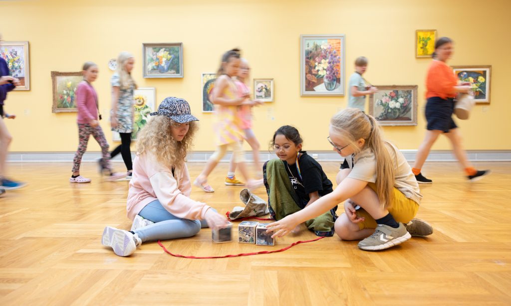 Kolme tyttöä istuu taidemuseon salissa lattialla keskittyneenä taidepajan tehtävään