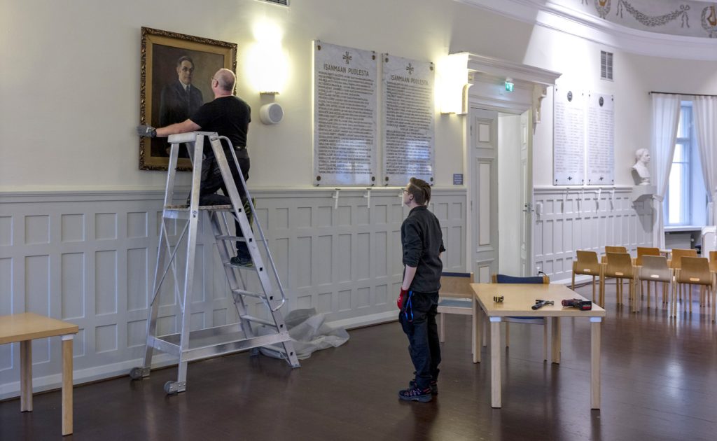 Museomestari ripustavaa suurikokoista taulua seinälle tikapuilla seisoen