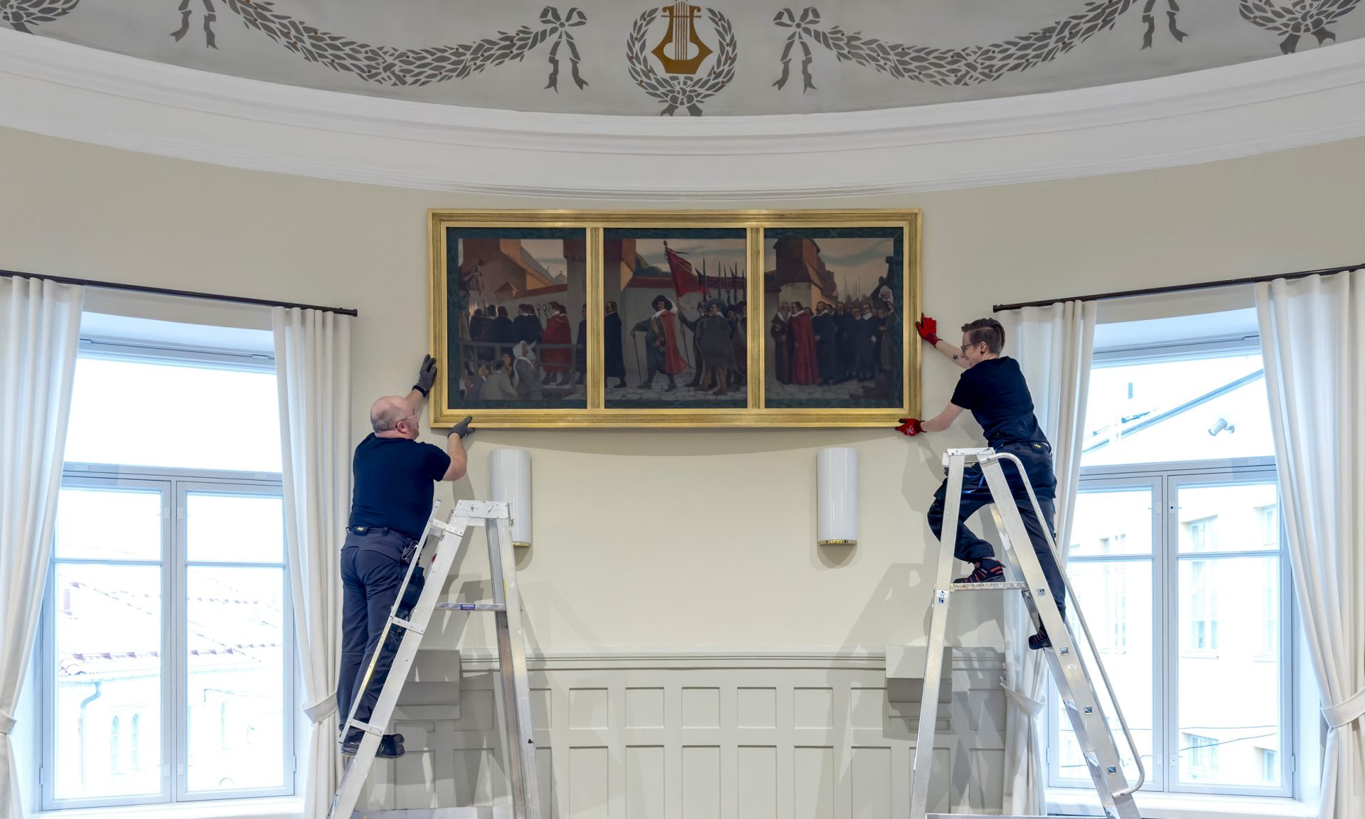 Exhibition technicians hang a large painting on the wall. They are standing on a ladder.