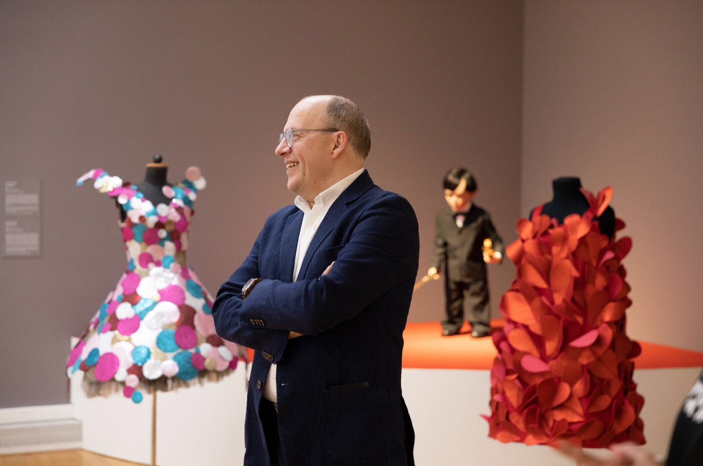 A smiling person stands with their arms crossed and looks to the left, behind them are clothes on mannequins.