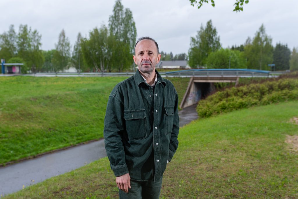 Henkilö seisoo toinen käsi taskussaan ja katselee kameraan. Taustalla vehreää luontoa ja silta.